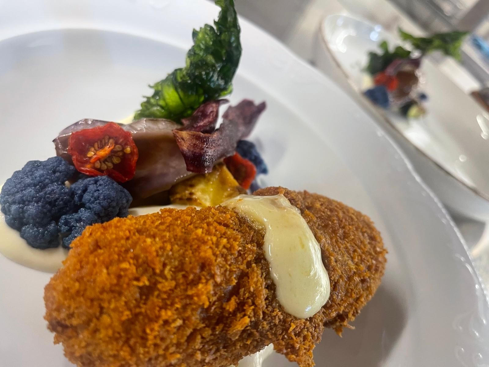 Eine goldbraun panierte Krokette mit heller Sauce, violettem Blumenkohl und getrockneten Tomaten